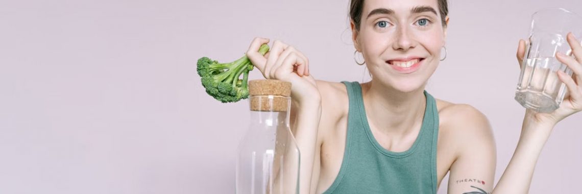 Woman in tank top smiling with broccoli, cabbage, and glass of water on table.